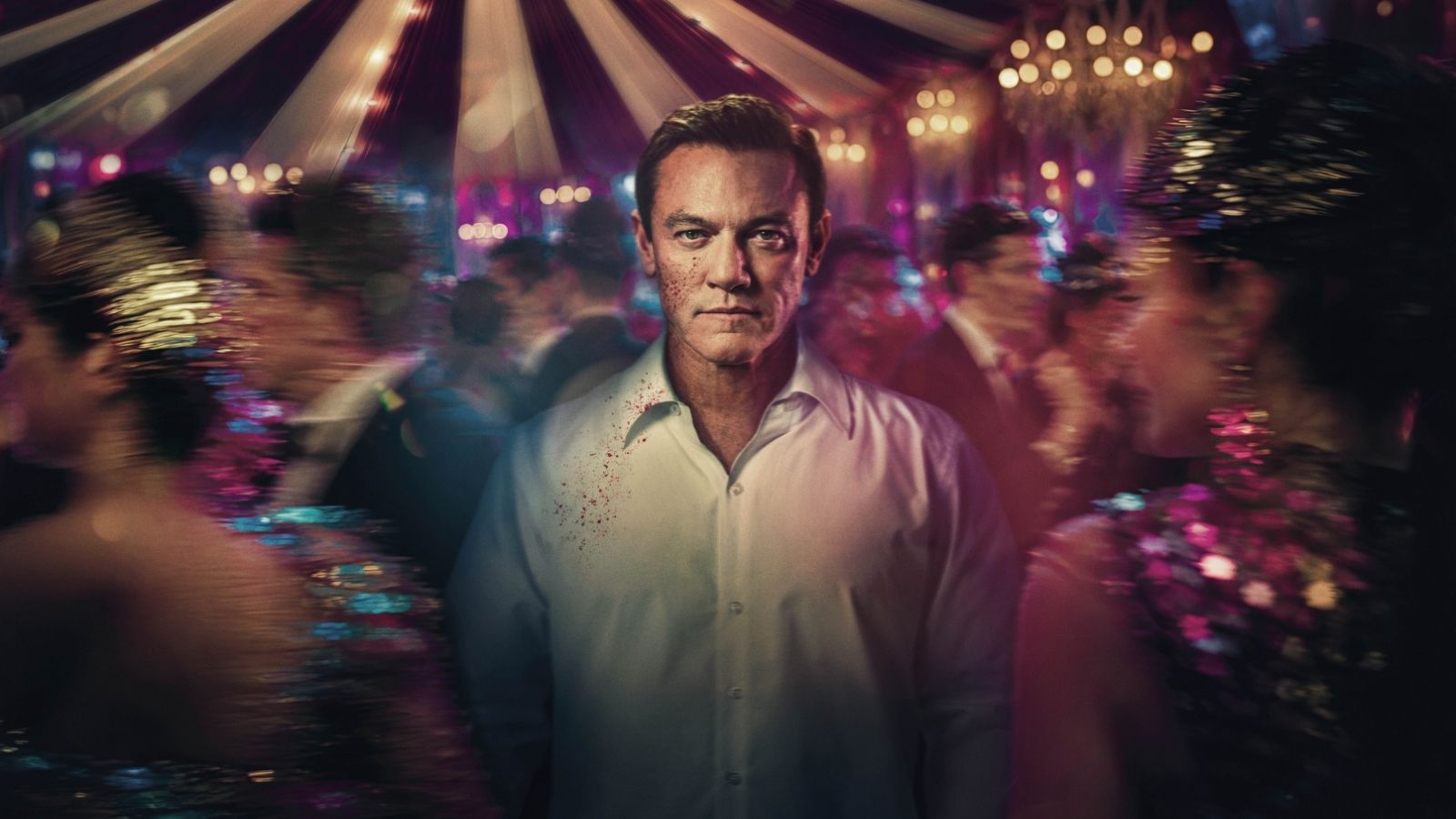 Luke Evans stood in the middle of a crowded scene with a circus tent in the background in the drama The Party