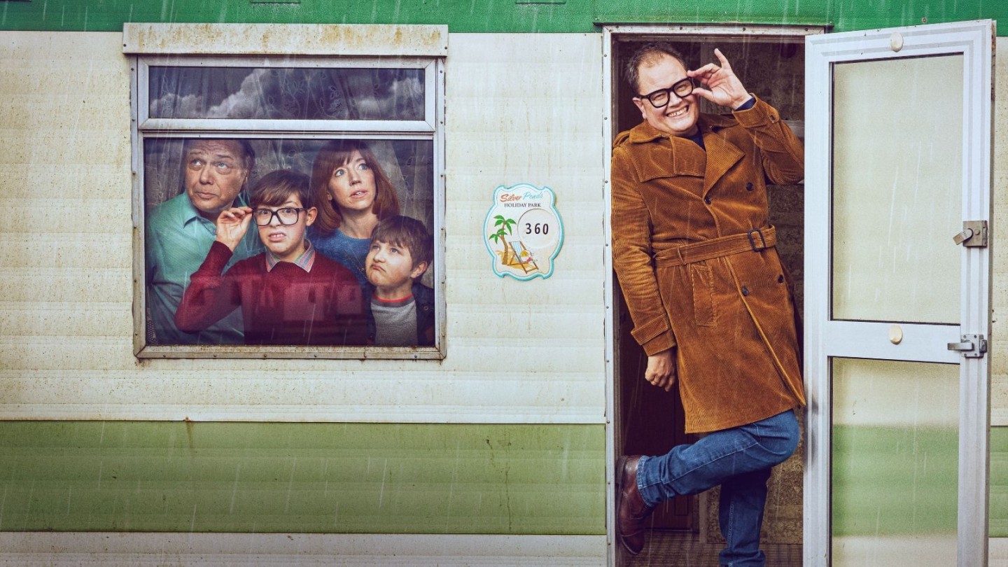  Promotional image for the TV series Changing Ends. On the right, Alan Carr as himself stands in the doorway of a white and green static caravan, wearing a mustard-coloured corduroy trench coat and thick-rimmed glasses, smiling and gesturing towards his face