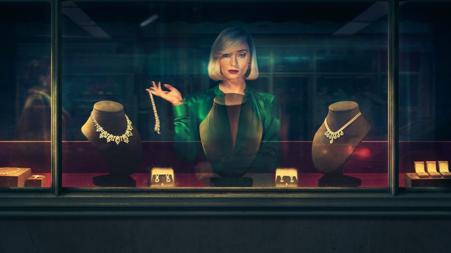 Promotional image for the TV series Joan. Sophie Turner as Joan Hannington stands behind a jewellery shop display window, looking directly at the camera. She wears a vibrant emerald green silk blouse and has a sleek, platinum blonde bob. Inside the brightly lit shop window, she holds up a diamond bracelet next to several ornate necklaces on velvet busts