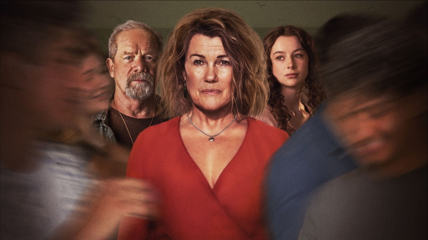 A promotional image for the TV series Happy Valley. At the centre is Sarah Lancashire as Catherine Cawood, wearing a burnt orange V-neck top and a heart-shaped necklace. She has a somber expression, and the background is dark and blurry, with ethereal, overlapping reflections of her face