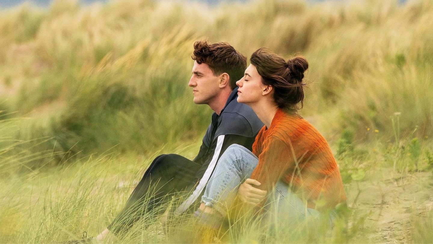 Two people sit close together on grassy sand dunes, dressed in casual clothing. One wears a dark jacket with striped sleeves, and the other wears a burnt orange jumper and light blue jeans. The setting is a natural coastal landscape with tall grass and soft, muted colours, suggesting a calm, outdoor scene.