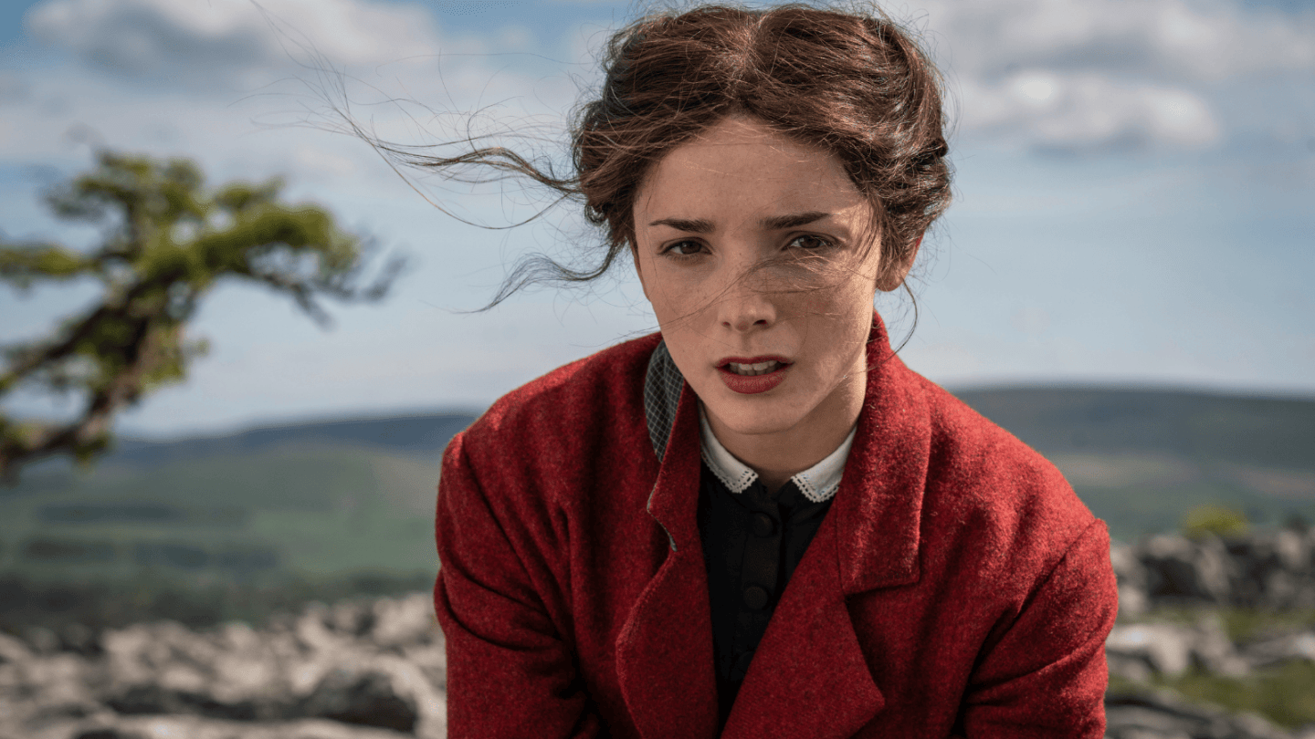 A person wearing a red coat sits outdoors on a rocky landscape with rolling hills and a partly cloudy sky in the background. Loose strands of hair blow in the wind. 