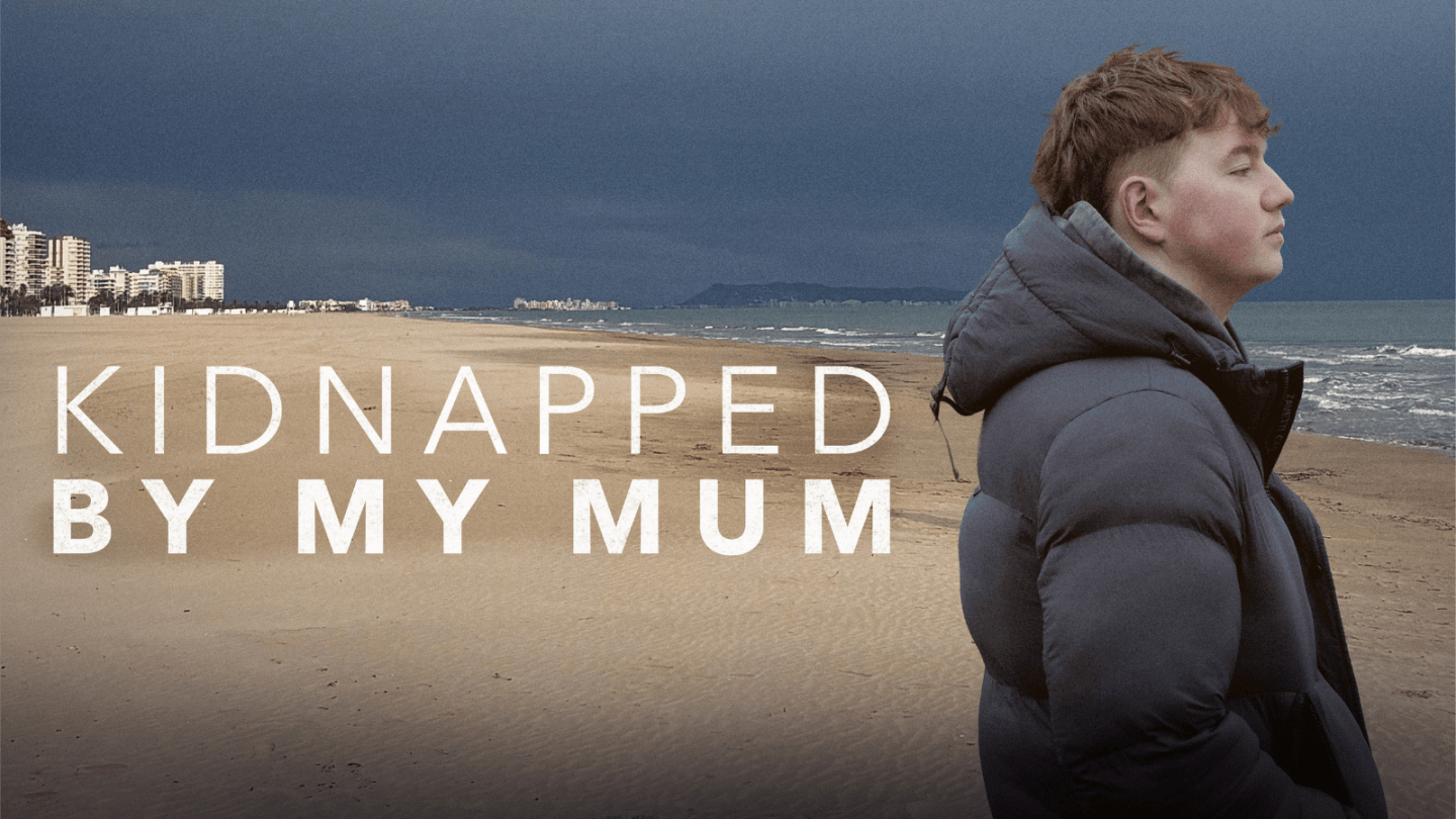 A Young Man wearing a puffer jacket stood on a beach with writing that reads kidnapped by my mum