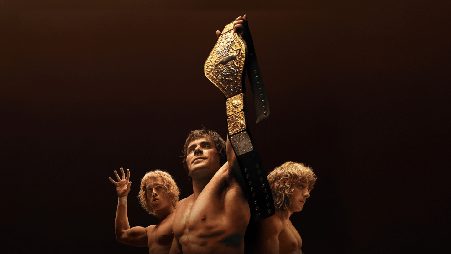 A man holding a golden wrestling belt with two young blonde men behind him against a dark background