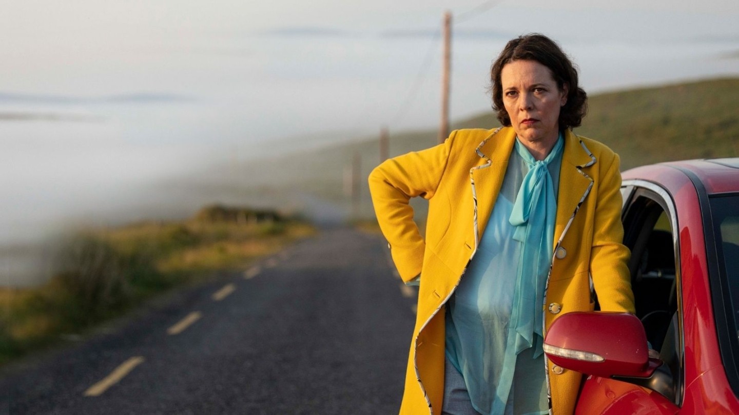 Woman wearing a yellow jacket with a blue dress and visible pregnancy bump stood next to a red car on a road by a cliff.