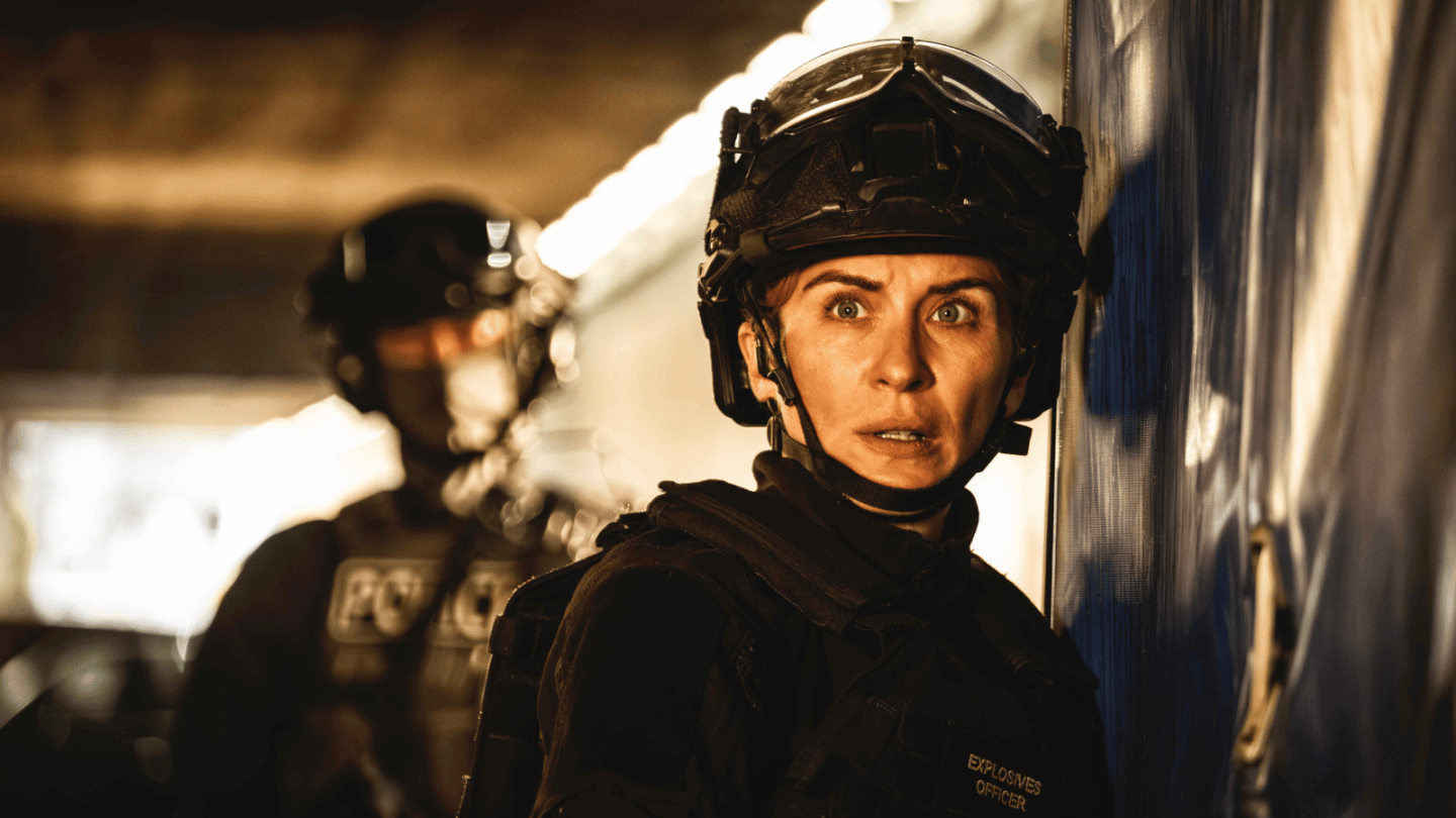 A tactical officer in protective gear stands beside a large vehicle or structure in a dimly lit environment, while another similarly equipped officer is visible out of focus behind. Strong artificial lighting creates bright highlights and deep shadows across the scene.