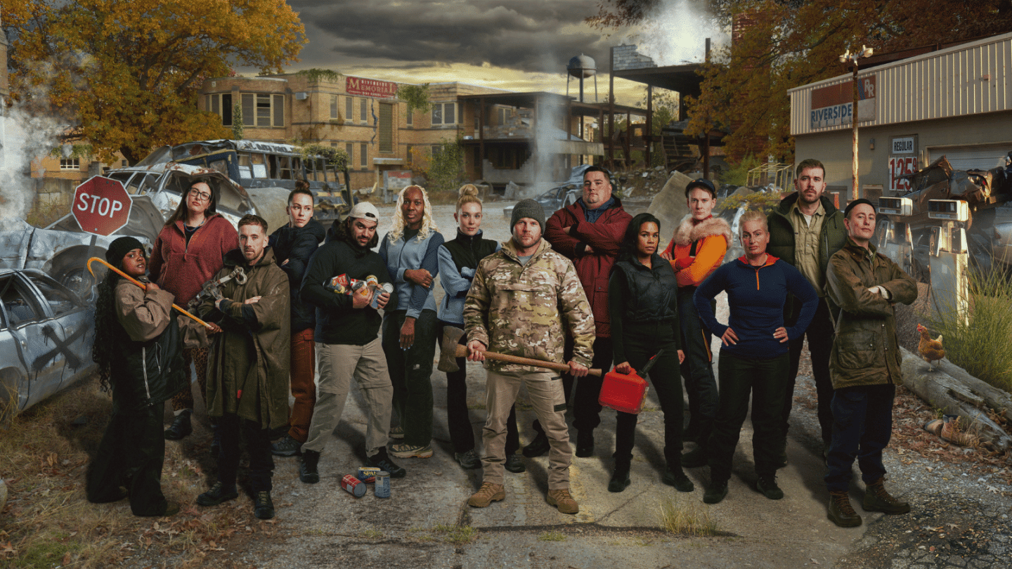 A group of contestants stands together in a post-apocalyptic town, surrounded by abandoned vehicles, damaged buildings and drifting smoke. The contestants are dressed in survival-style clothing and hold items including tools, fuel cans and supplies.
