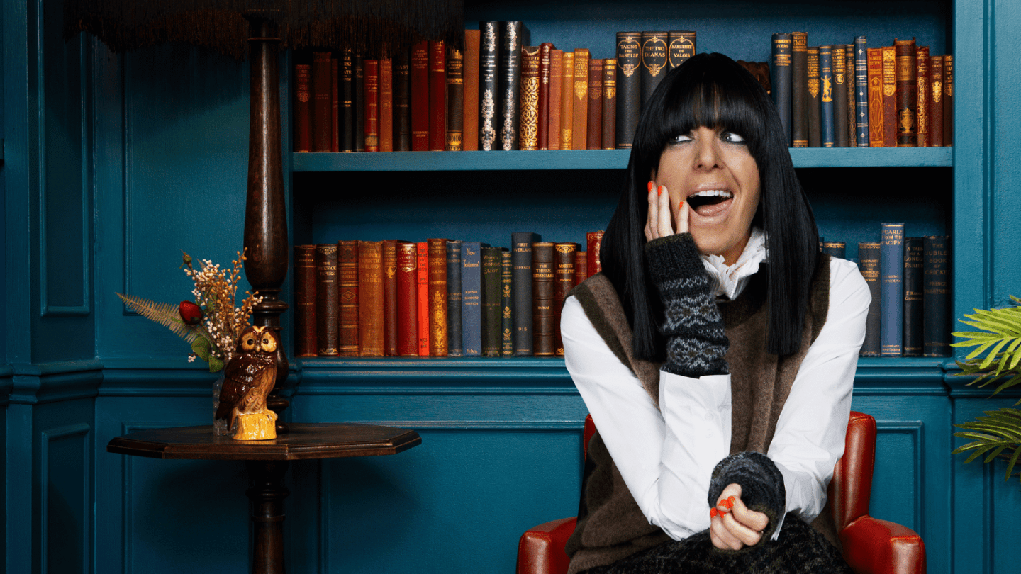 A person with a dark fringe sits in front of a bookshelf filled with vintage books, resting a hand against their face. The room features deep blue walls, a small owl ornament on a side table, and warm, cosy lighting.