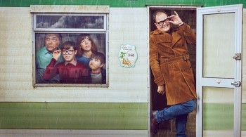  Promotional image for the TV series Changing Ends. On the right, Alan Carr as himself stands in the doorway of a white and green static caravan, wearing a mustard-coloured corduroy trench coat and thick-rimmed glasses, smiling and gesturing towards his face