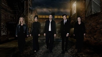 Six people walk towards the camera along a dark, cobbled street at night, surrounded by brick buildings and streetlights, with a railway bridge behind them.
