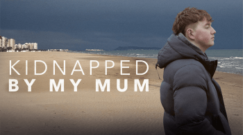 A Young Man wearing a puffer jacket stood on a beach with writing that reads kidnapped by my mum
