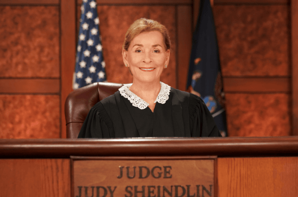 A judge wearing a black robe with a white lace collar sits at a courtroom bench. Behind the judge are wooden panels and two flags. The front of the bench displays a nameplate reading ‘Judge Judy Sheindlin.