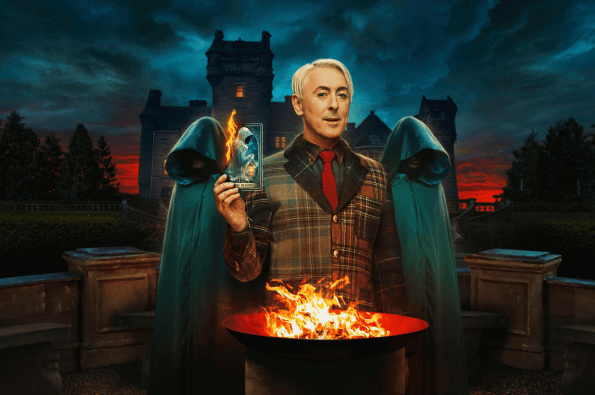 Alan Cumming dressed in a checked suit holding a tarot card with two hooded traitors stood behind him. An old gothic house in the background.