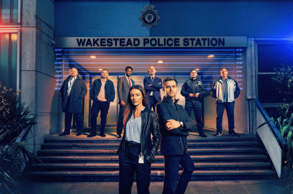 Promotional cast image for a crime drama showing a group of detectives and police officers standing outside Wakestead Police Station at night. Two lead characters stand in the foreground, while the wider team gathers on the station steps behind them, illuminated by blue and red police lights.