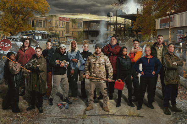 A group of contestants stands together in a post-apocalyptic town, surrounded by abandoned vehicles, damaged buildings and drifting smoke. The contestants are dressed in survival-style clothing and hold items including tools, fuel cans and supplies.