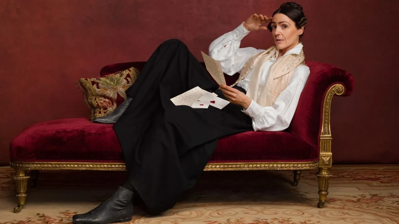 Promotional image for the TV series Gentleman Jack. Suranne Jones as Anne Lister is reclining on a luxurious red velvet chaise longue against a dark red wall.