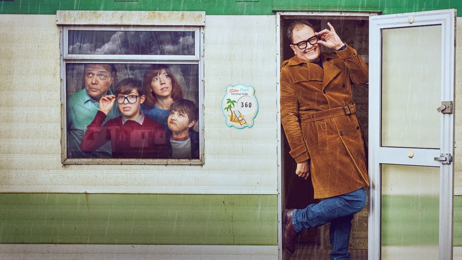  Promotional image for the TV series Changing Ends. On the right, Alan Carr as himself stands in the doorway of a white and green static caravan, wearing a mustard-coloured corduroy trench coat and thick-rimmed glasses, smiling and gesturing towards his face