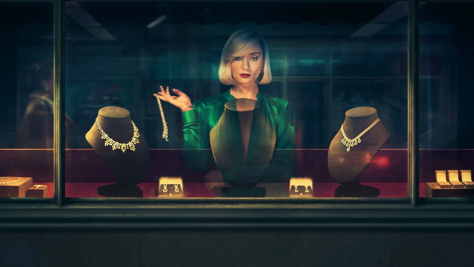 Promotional image for the TV series Joan. Sophie Turner as Joan Hannington stands behind a jewellery shop display window, looking directly at the camera. She wears a vibrant emerald green silk blouse and has a sleek, platinum blonde bob. Inside the brightly lit shop window, she holds up a diamond bracelet next to several ornate necklaces on velvet busts