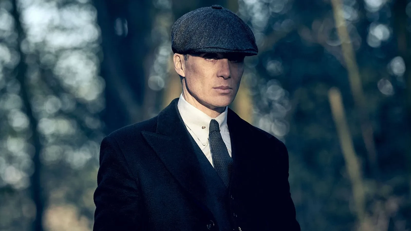 A close-up of Cillian Murphy as Thomas "Tommy" Shelby, the leader of the Peaky Blinders gang. He is wearing a dark, heavy overcoat over a three-piece suit and a distinctive tweed flat cap