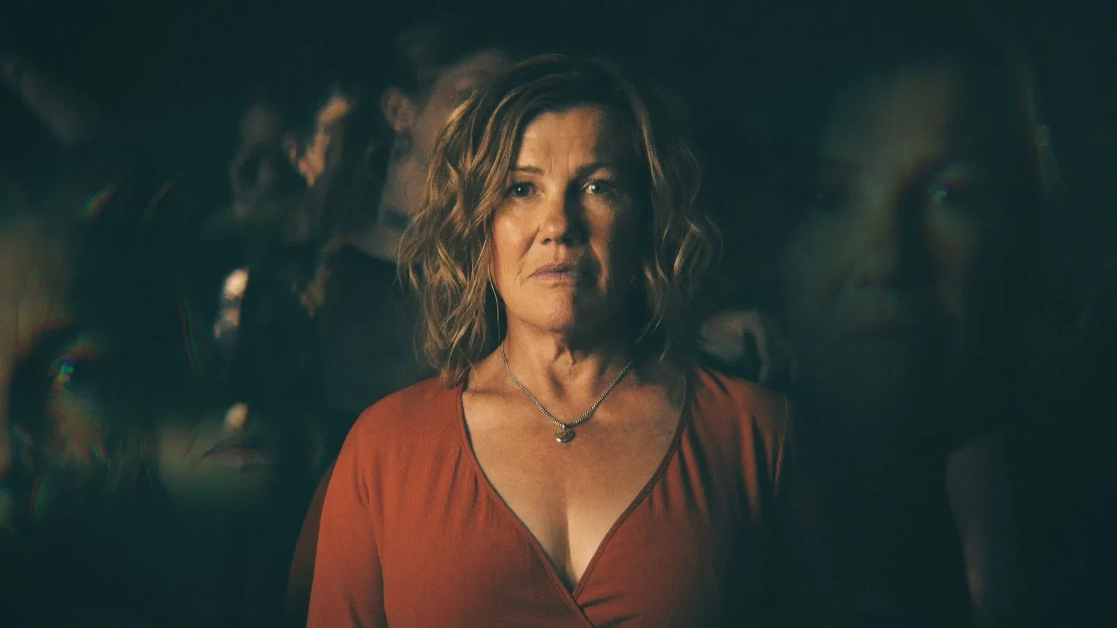 A promotional image for the TV series After the Party. Robyn Malcolm as Penny Wilding stands in the centre wearing a burnt orange V-neck top and a heart-shaped necklace. She has a somber expression, set against a dark, blurry background with ethereal, overlapping reflections of her face.
