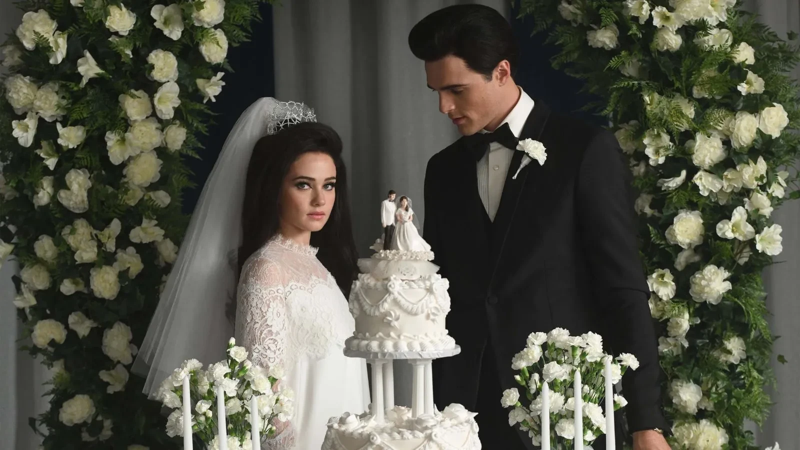 A women ina wedding dress and veil next to a wedding cake and her husband in a suit. With a rose arch behind them.
