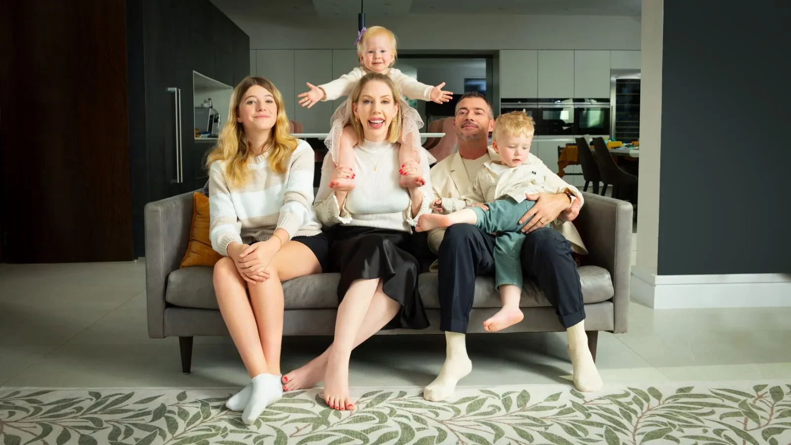 A man and women with two young kids and one teenager sat on a grey sofa in a living room 