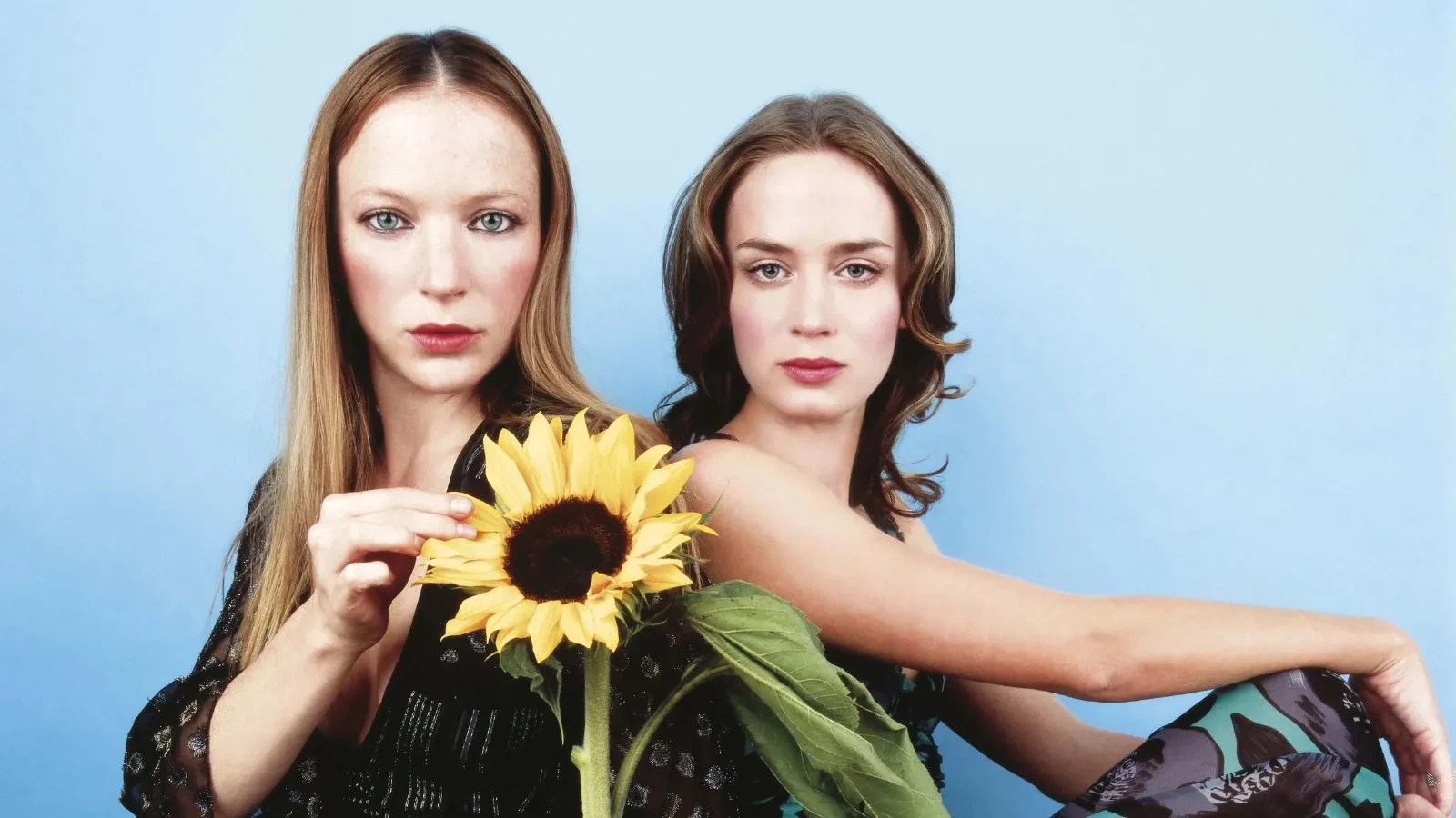 A blonde women holding a sunflower looking into the camera, a brunette women at next to her staring