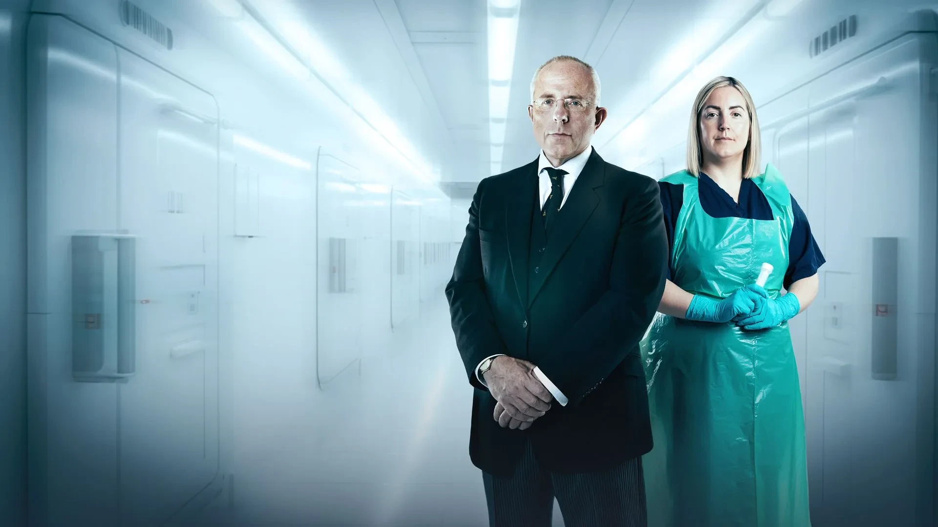 A man in a suit stood next to a women in a blue plastic apron stood in a hospital