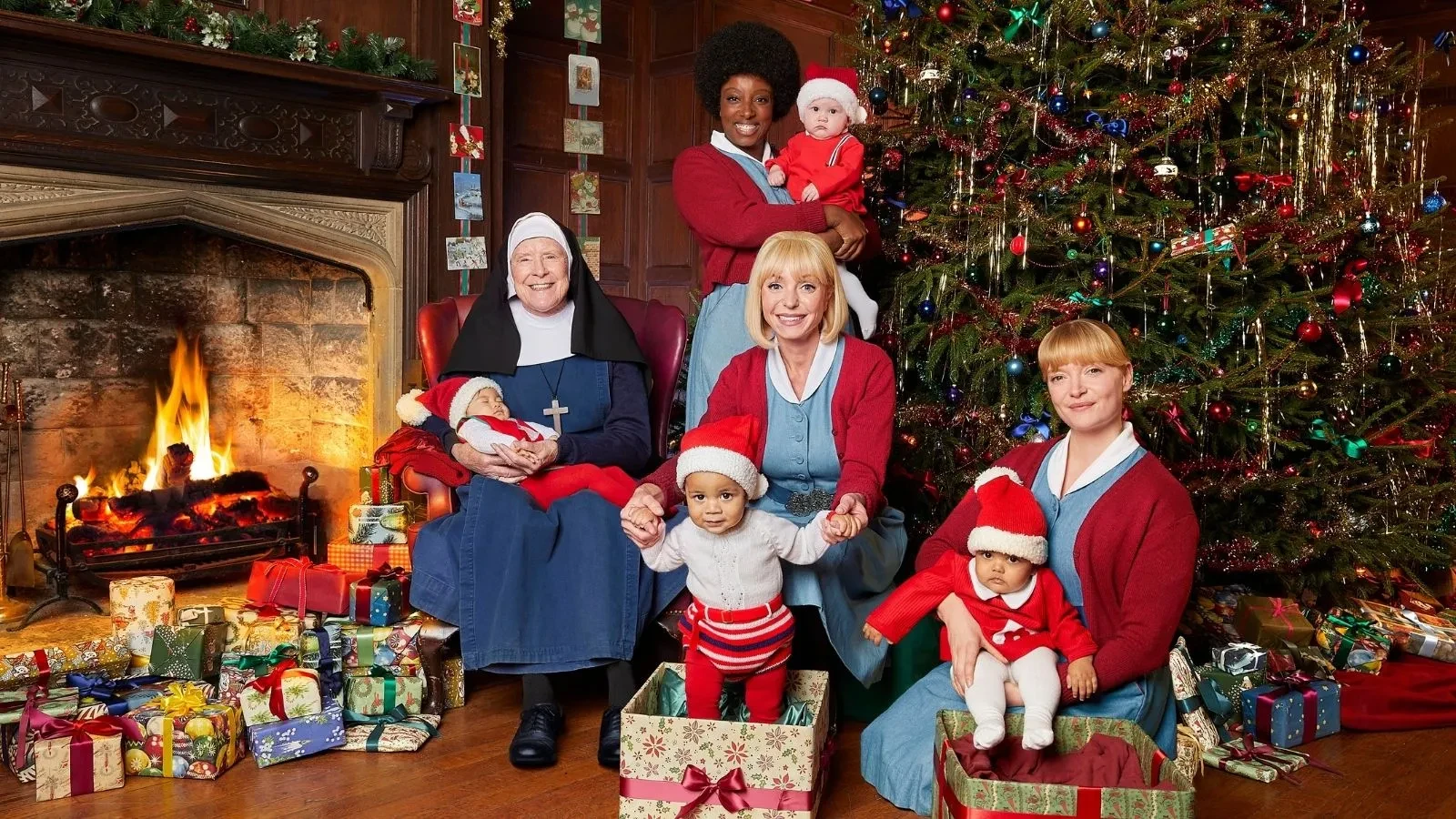 Group sitting and standing near a large Christmas tree and a fireplace, surrounded by wrapped gifts and holiday decorations.