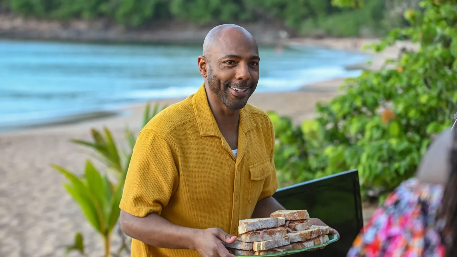 Person wearing a yellow short-sleeve shirt holding a tray of stacked sandwiches on a tropical beach with greenery and ocean in the background.