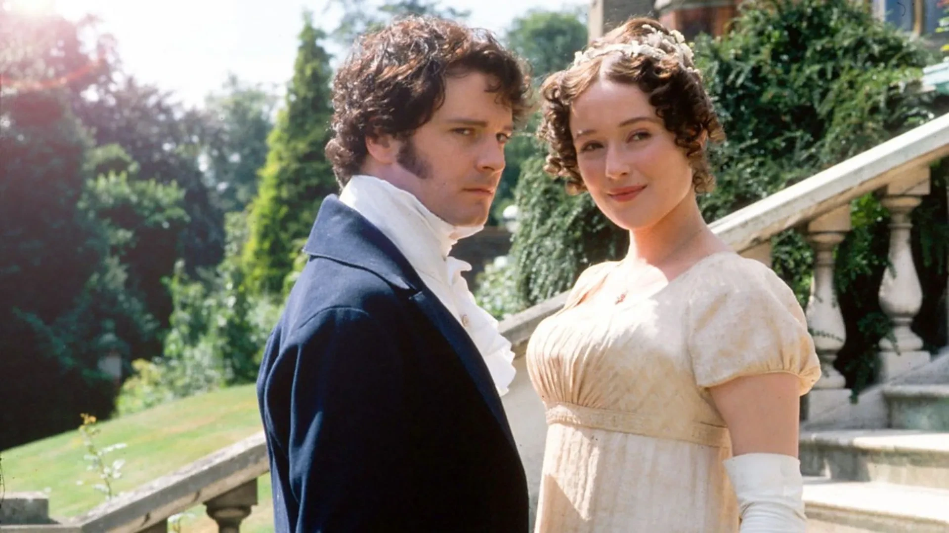 Still image from the 1995 TV series Pride & Prejudice showing a man and woman in Regency-era dress standing on stone steps outdoors, with trees and a country garden behind them.