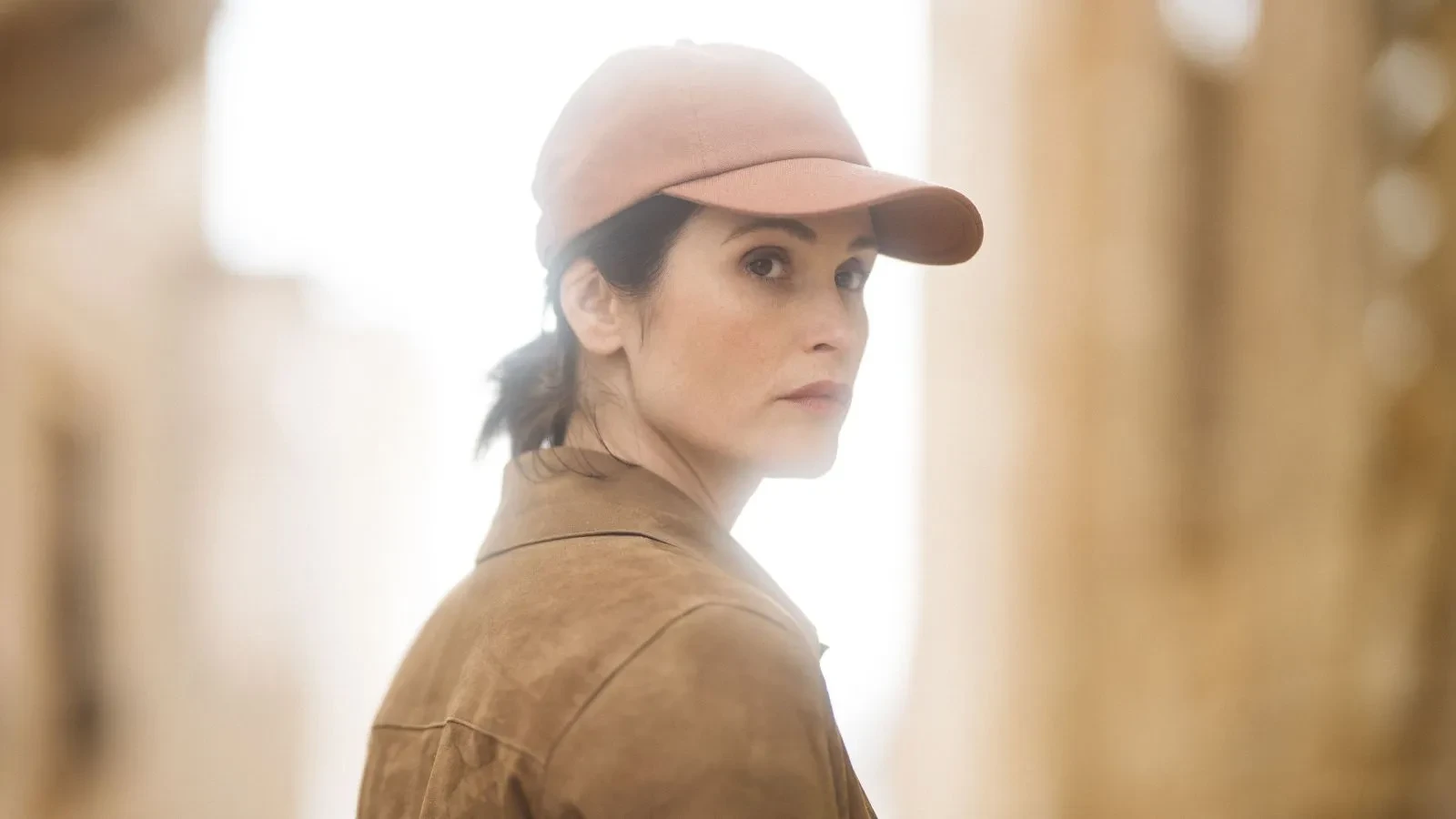 A person wearing a brown jacket and a matching cap stands outdoors in a sunlit, sandy environment, looking over their shoulder. The background is softly blurred, suggesting an arid or desert-like location.