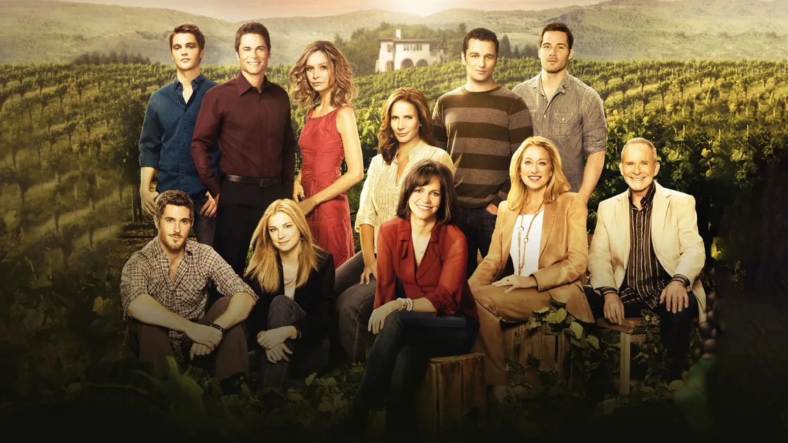 A large family group posing together in a vineyard at sunset, with rolling hills and a farmhouse in the background. They are dressed in a mix of smart‑casual and formal clothing, creating a warm, atmospheric scene that suggests themes of family, relationships and rural lifestyle.