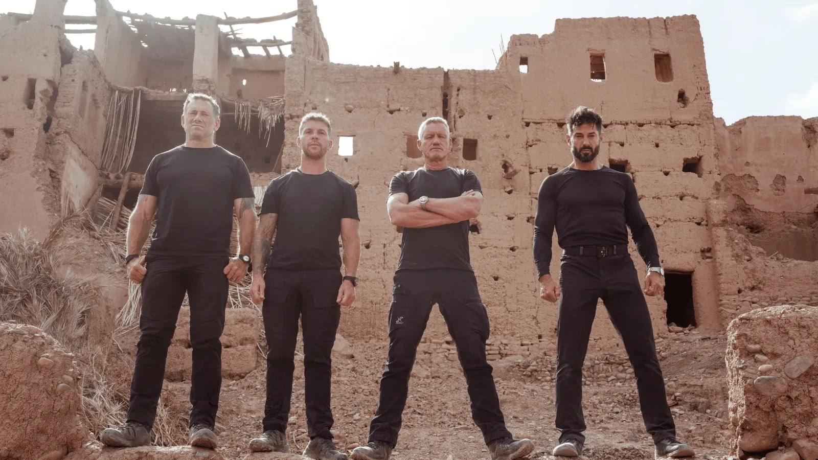 Four individuals dressed in black tactical-style clothing stand in front of a crumbling, sand-coloured building, posing confidently in a rugged outdoor environment.