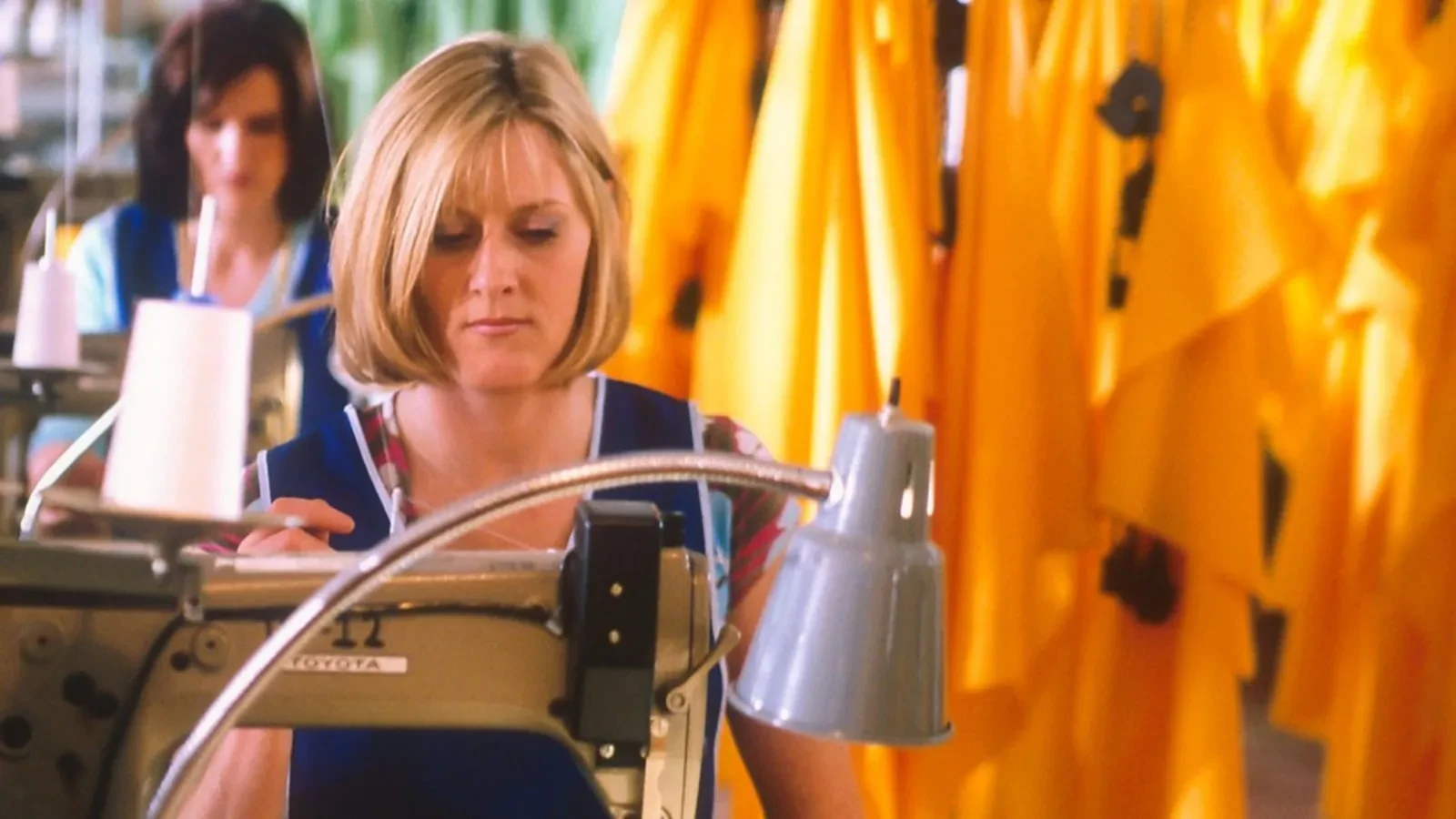 A textile factory worker sits at a sewing machine, stitching fabric under a task lamp, with bright yellow garments hanging in rows behind her and another worker visible in the background, creating a busy production‑line atmosphere.
