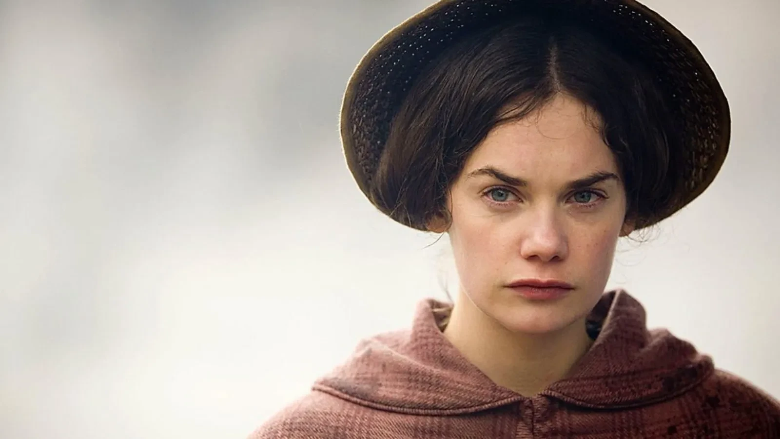 Close-up of a person wearing a brown bonnet and a textured pink cloak with a rounded collar, set against a soft, light background. The clothing style suggests a historical or period setting.