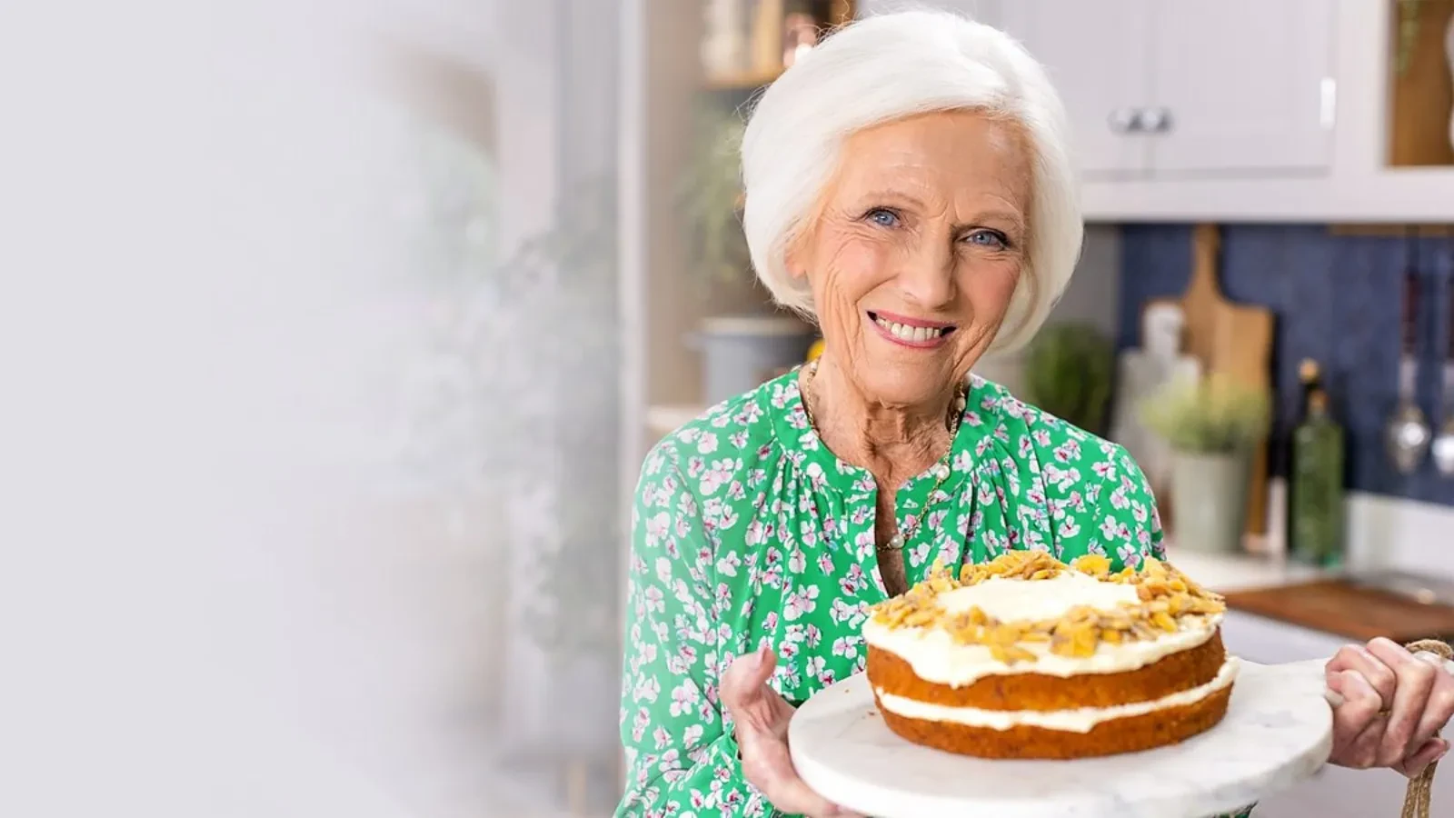 An older woman in a green floral blouse holding a homemade layered carrot cake topped with nuts on a marble cake stand, standing in a bright, modern kitchen setting.