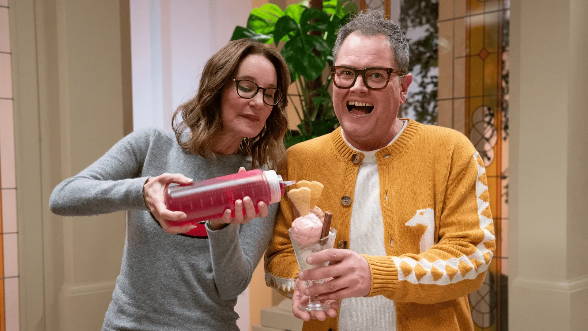 Alan Carr in a yellow cardigan with ducks on holding a sundae with Susie Dent wearing a grey long sleeved top pouring red sauce onto the sunday