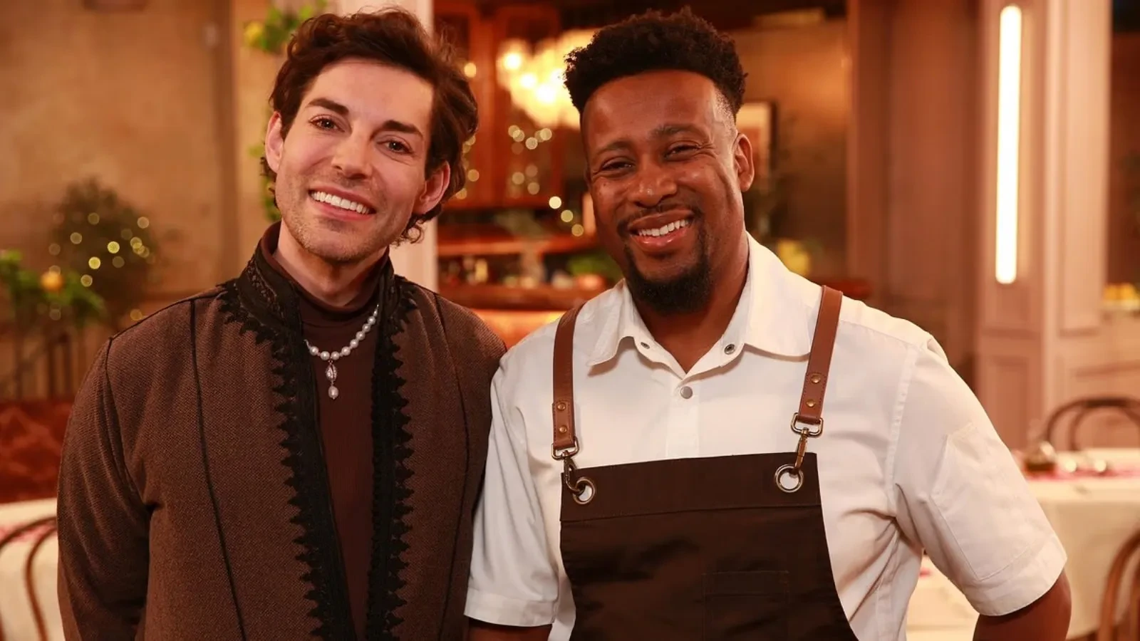 Two people standing inside a warmly lit restaurant, one wearing a brown patterned jacket with a pearl necklace and the other dressed in a white shirt with a brown apron, surrounded by soft ambient lighting and elegant dining décor.