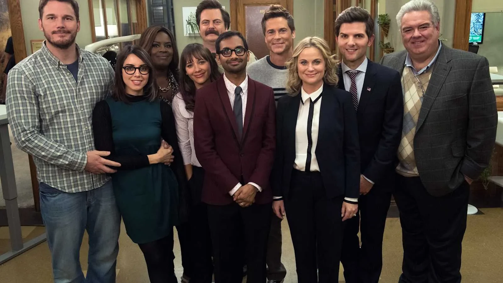 A group of people stand closely together inside a government‑office setting, dressed in a mix of business attire and smart‑casual clothing. The group is arranged in a friendly, posed formation, with wooden doors, office décor, and soft lighting creating a warm workplace atmosphere in the background