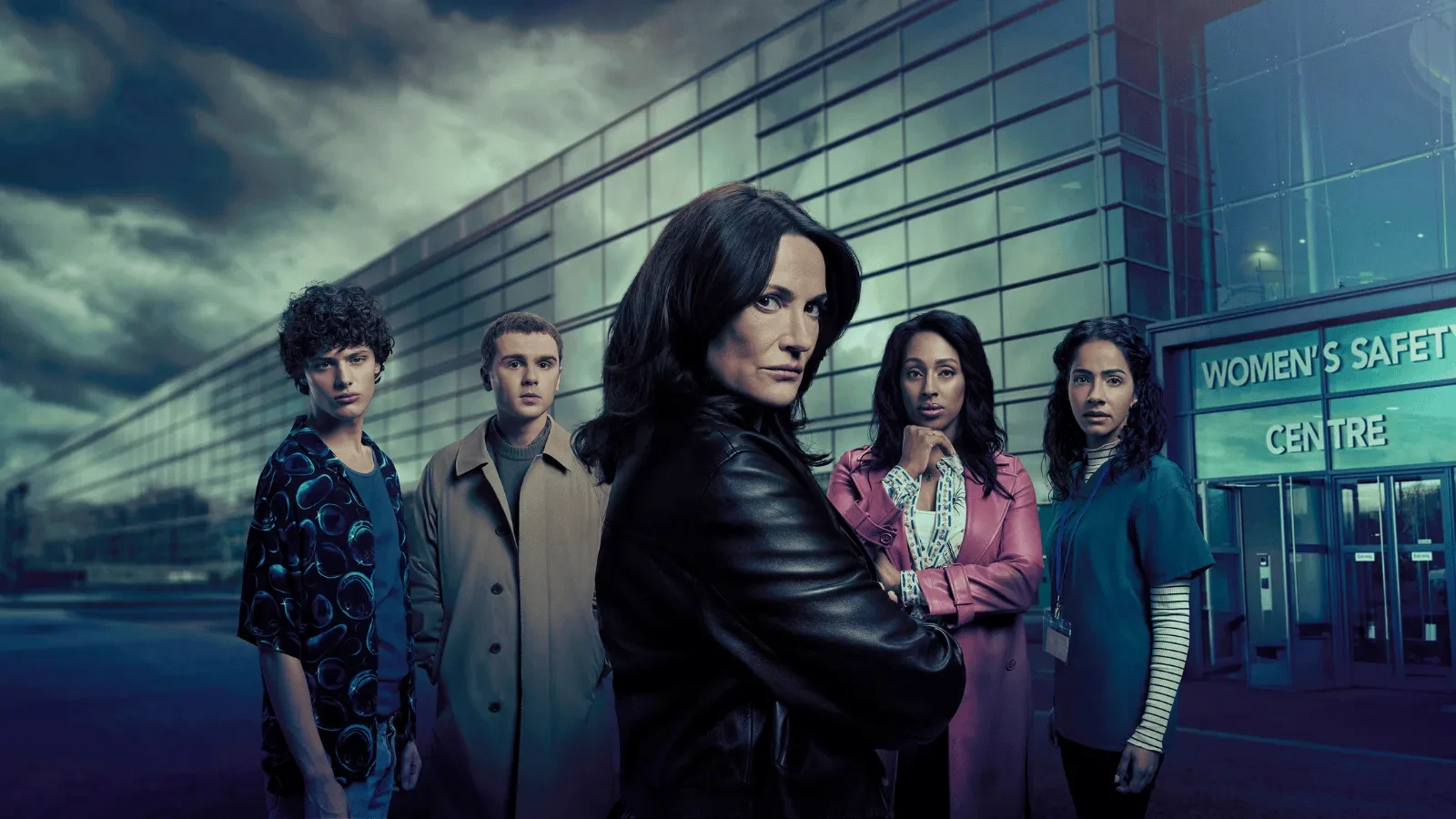 A group of people standing together outside a modern glass building marked ‘Women’s Safety Centre,’ set against a dark, atmospheric sky.