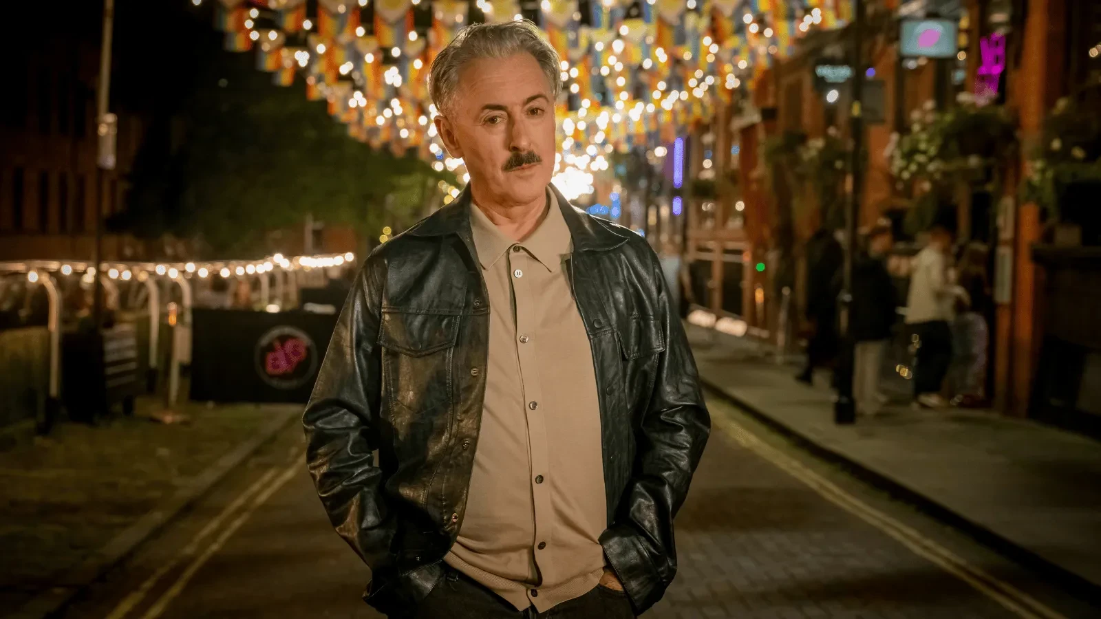Man wearing a leather jacket standing in a brightly lit street at night, surrounded by hanging lanterns and city nightlife