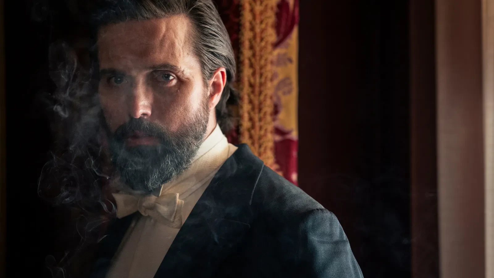 A person in formal vintage attire, including a bow tie and dark suit jacket, stands indoors near ornate curtains, with smoke drifting through the scene. 
