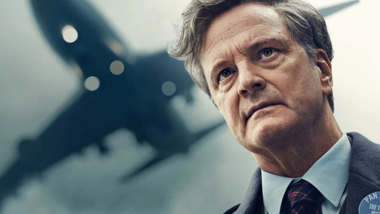 Person in a suit standing outdoors with a large airplane flying low in the background.