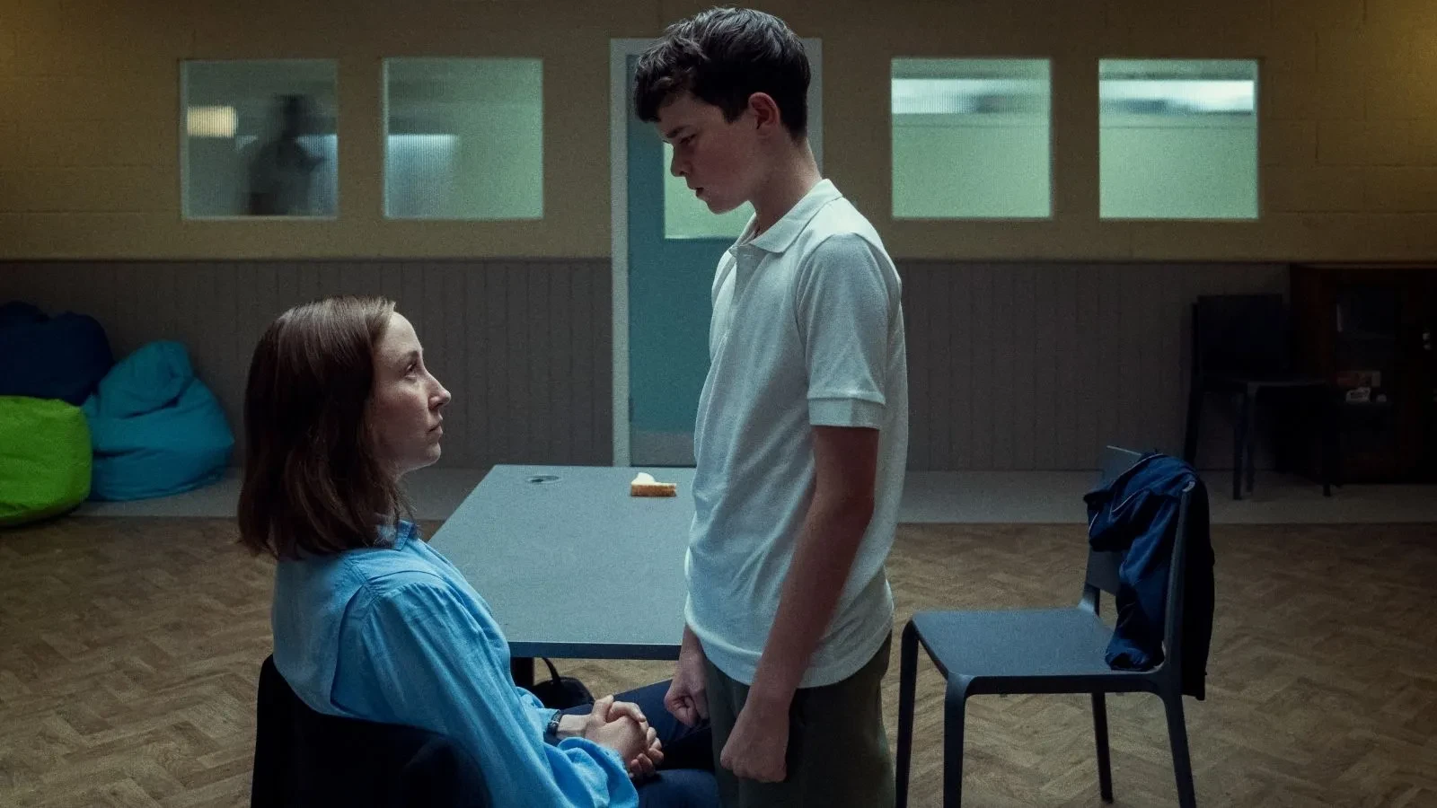 Two people in a meeting room, one seated and one standing, holding hands across a table in a calm, quiet environment.
