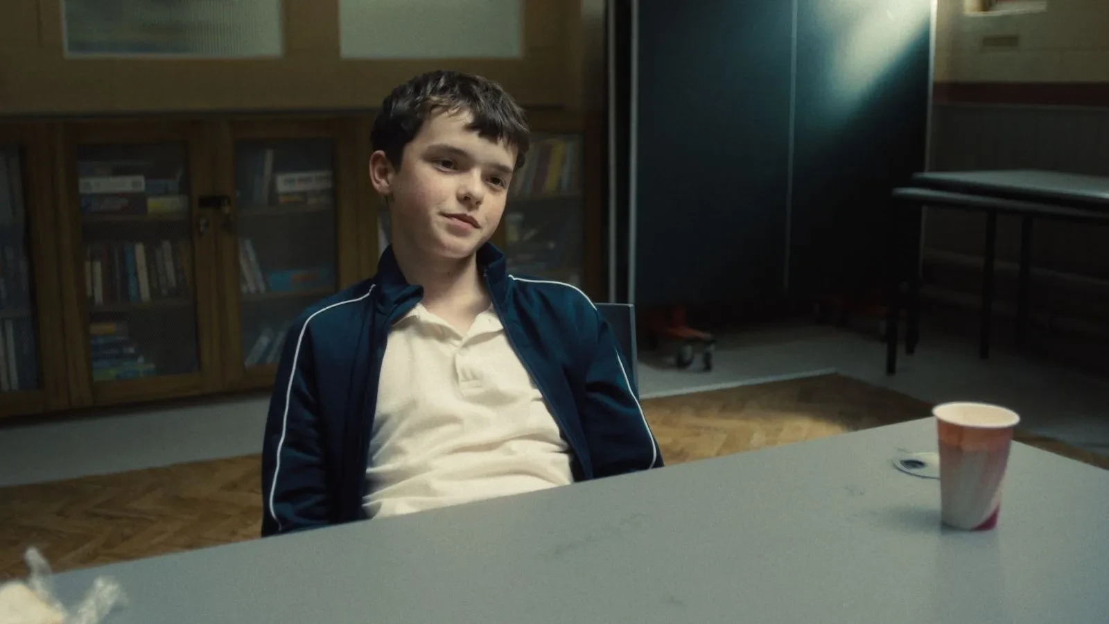 Individual in a tracksuit sitting at a table in a dim community room with bookshelves and a paper cup nearby.