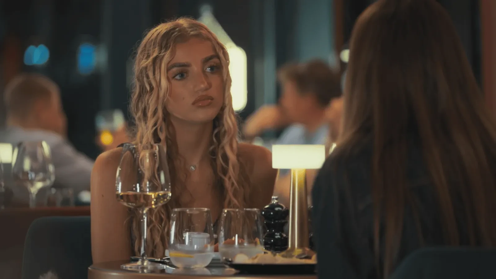 A person with long wavy hair sits at a dimly lit restaurant table with wine glasses, plates, and a small glowing table lamp, speaking to another person seated opposite. 