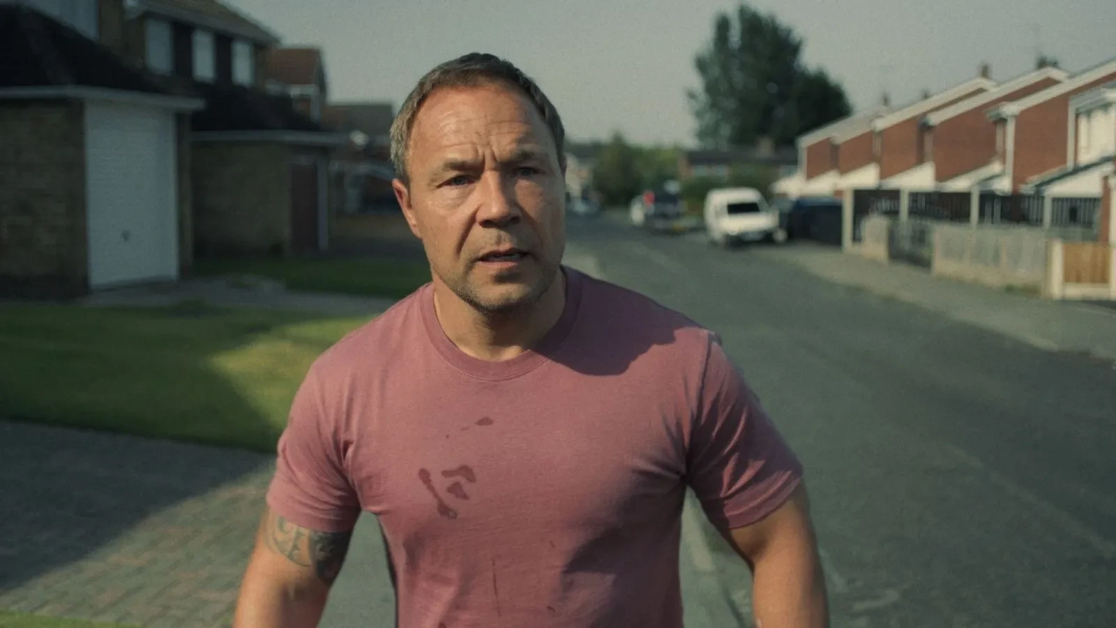 Person in a pink T‑shirt walking down a residential street lined with houses on a sunny day.