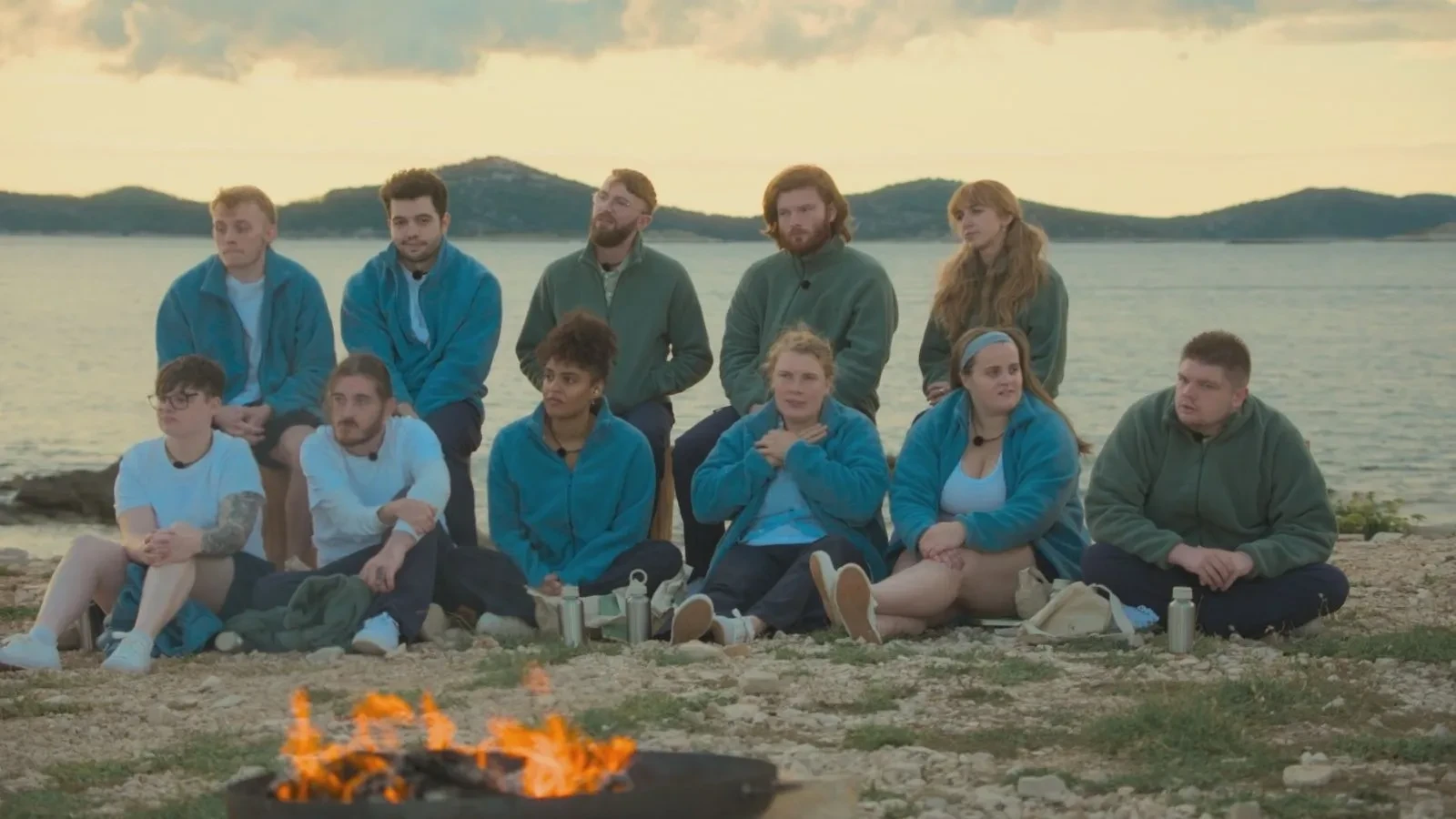 A large group of people seated on rocky ground beside the sea at dusk, wearing matching outdoor clothing and facing a campfire with hills and water in the background.