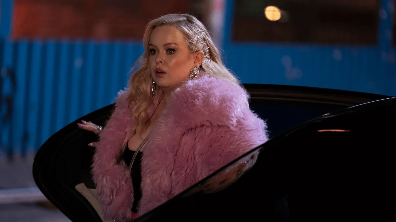 A person stepping out of a car at night wearing a fluffy pink coat, photographed in a street setting with blue fencing behind them. 